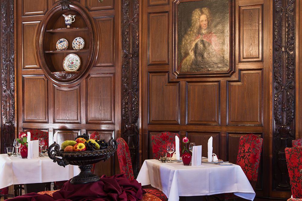 Elegantes Restaurant mit holzgetäfelten Wänden, einem Gemälde und dekorativen Tellern. Perfekt für Feste, die Tische sind mit weißen Tischdecken und roten Stühlen gedeckt, auf einem Tisch steht ein Obstgedeck, das eine einladende Esszimmeratmosphäre schafft.