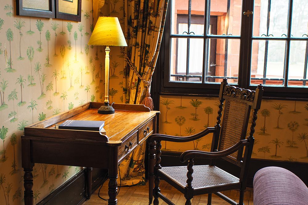 A wooden desk with a book and lamp sits next to an upholstered chair in the cozy conservatory. The room features palm-themed wallpaper and a barred window covered with floral curtains, creating a sense of relaxation.