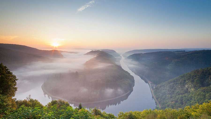 Der Sonnenaufgang über einer nebligen Flussbiegung, umgeben von üppigen, grünen Hügeln unter einem klaren Himmel, lädt zu einem Gefühl der Entspannung ein.