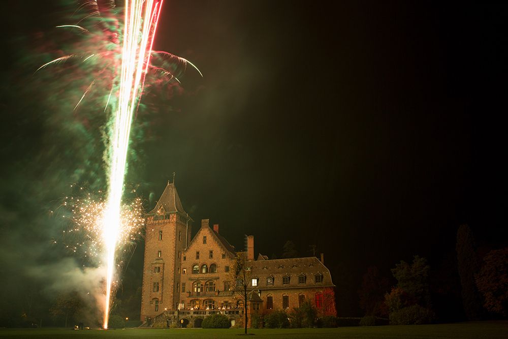 Fireworks light up the sky at night over a large, historic brick mansion surrounded by dark foliage.