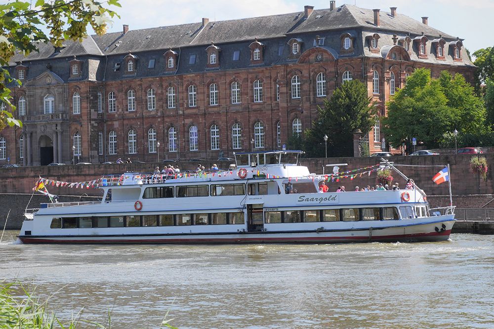 A white tour boat named “Saargold” cruises on a river, with a historic brick building in the background, perfect for those looking for day trips near their hotel.