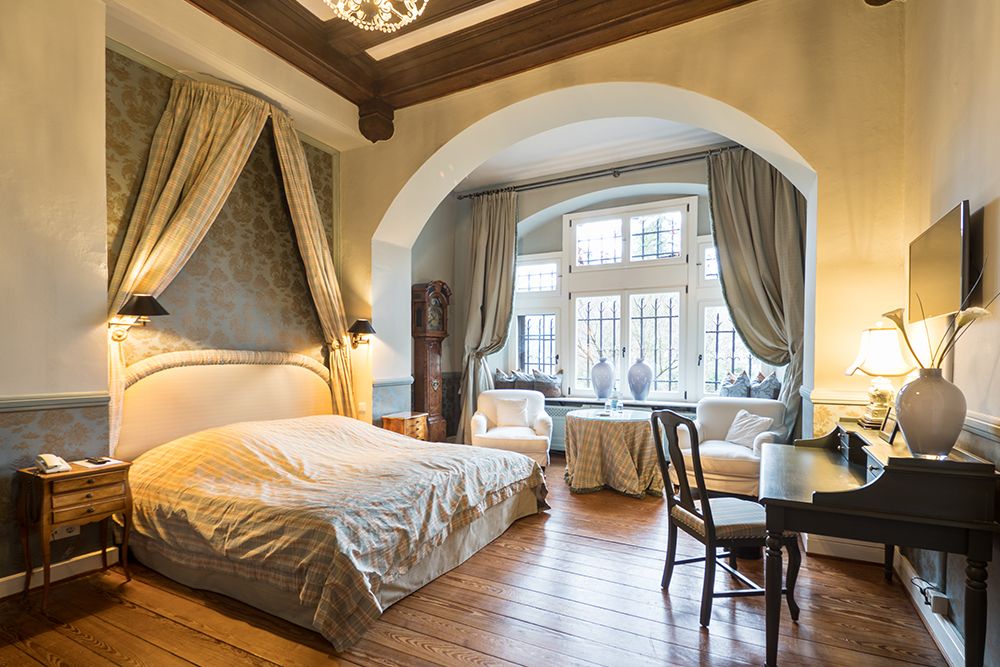 Elegant eingerichtetes Schlafzimmer mit Himmelbett, Holzboden, Schreibtisch, Stühlen und großen Fenstern mit Vorhängen.