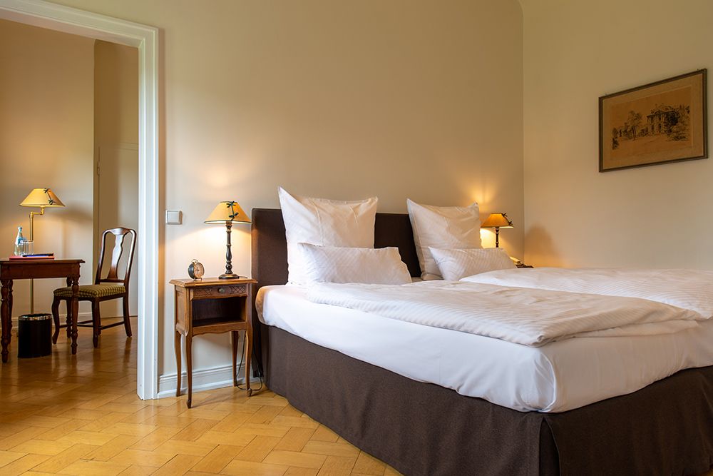 Ein ordentlich gemachtes Doppelbett mit weißen Kissen und Tagesdecke in einem warm beleuchteten Raum mit Holzboden und Beistelltisch.