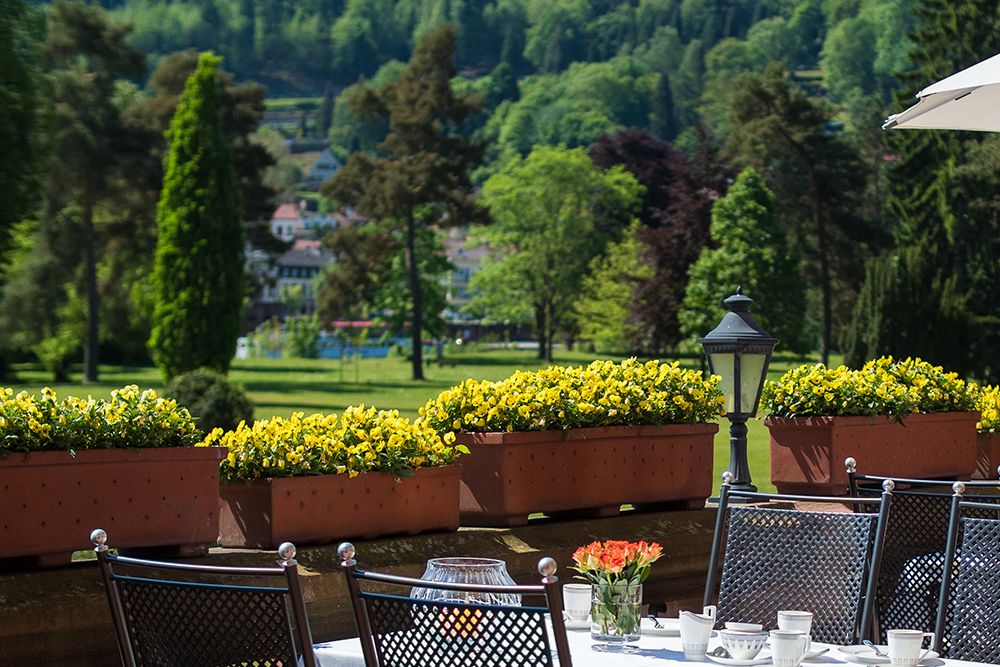 Essbereich im Freien mit Metallstühlen und Tischen mit Glasgeschirr, mit Blick auf Saars üppigen Garten. Gelbe Blumen säumen die Schlossterrasse, mit hohen Bäumen und entfernten Gebäuden im Hintergrund.