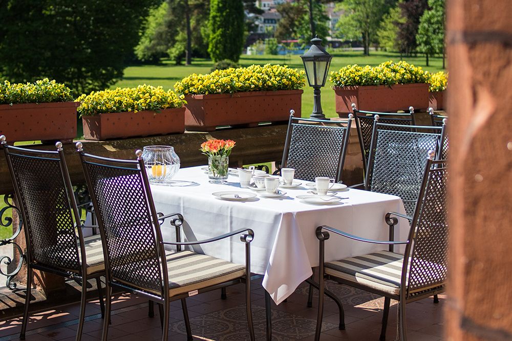Dieser Esstisch im Freien steht auf der bezaubernden Schlossterrasse und ist mit einem frischen weißen Tuch bedeckt, auf dem Tassen, Untertassen, ein Krug und eine Blumenvase stehen. Umgeben von Metallstühlen und eingebettet in das üppige Grün des Saarparks bietet er eine idyllische Kulisse für gemütliche Zusammenkünfte.