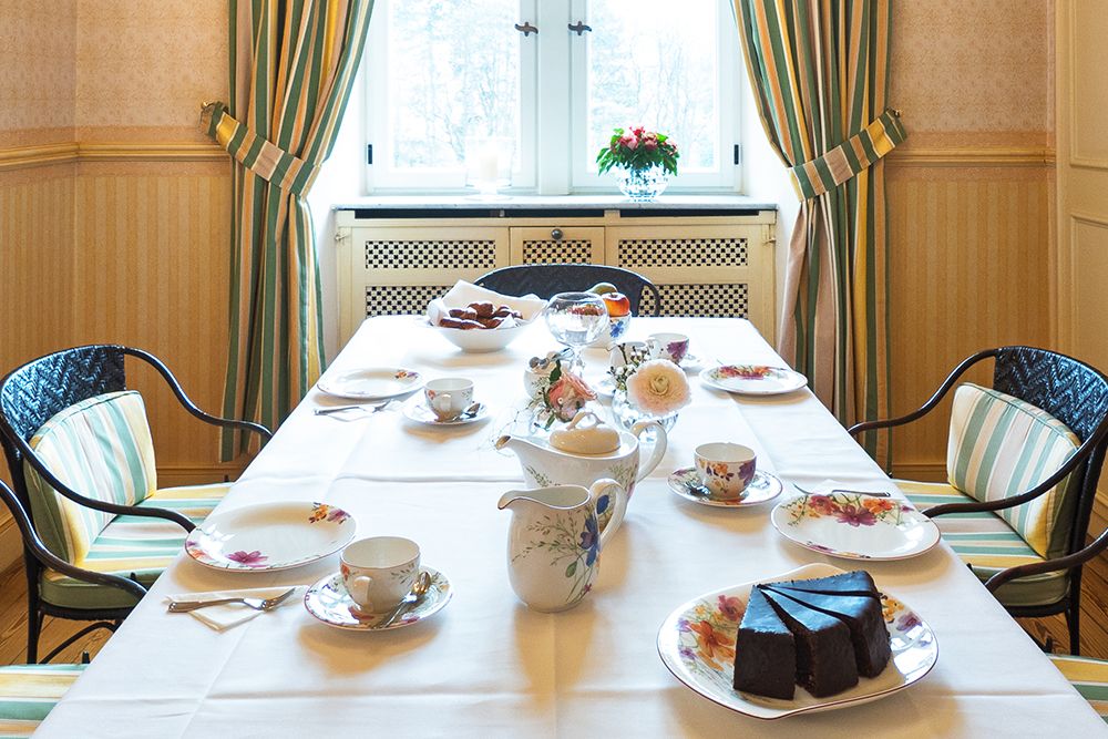 Der für die Teestunde gedeckte Esstisch besteht aus exquisitem Porzellan mit Blumenmotiven und einem reichhaltigen Schokoladenkuchen, alles eingerahmt von den gestreiften Vorhängen des Gelben Zimmers. Ein zartes Blumengesteck verleiht Charme und lädt Sie ein, das gemütliche Ambiente zu genießen, das an den bezaubernden Reiz des Saareckparks erinnert.