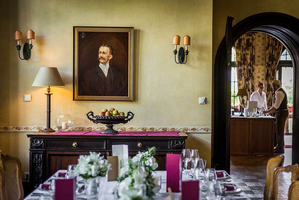 A dining room with a table set for a meal, with white flowers and red menus. A hallway in the background shows two people at a bar. A portrait and lamps hang on the wall above a sideboard.
