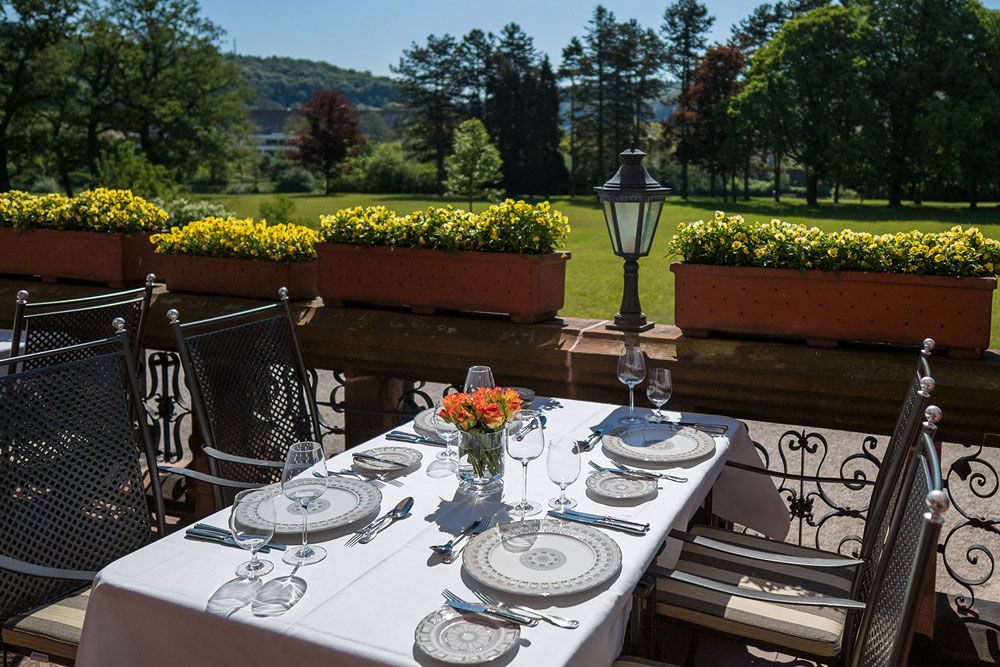Esstisch für vier Personen im Freien auf einer Terrasse mit Blick auf einen Garten mit Bäumen und Blumenkästen.