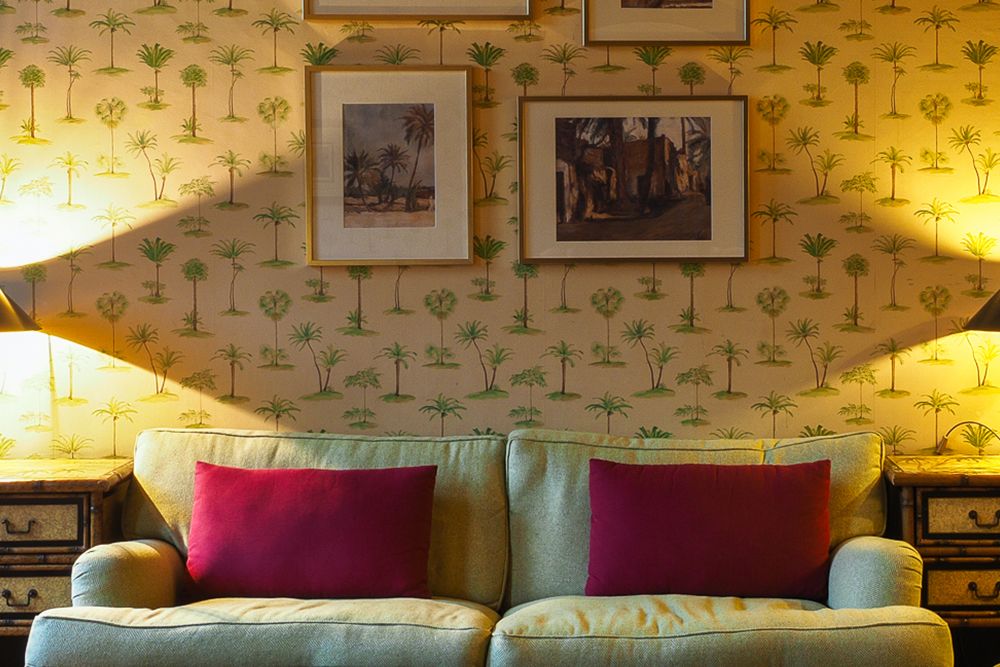 A green sofa with red cushions nestles against beige wallpaper with a palm pattern, evoking a conservatory feel. Framed paintings and two lamps decorate the wall above, while wooden side tables flank it, creating an oasis of relaxation.
