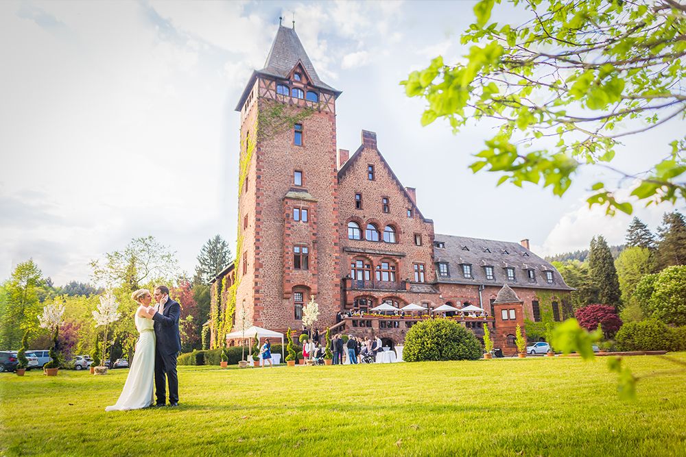 A couple in their wedding dresses pose in front of a large, historic brick building with a tower, surrounded by greenery and guests.