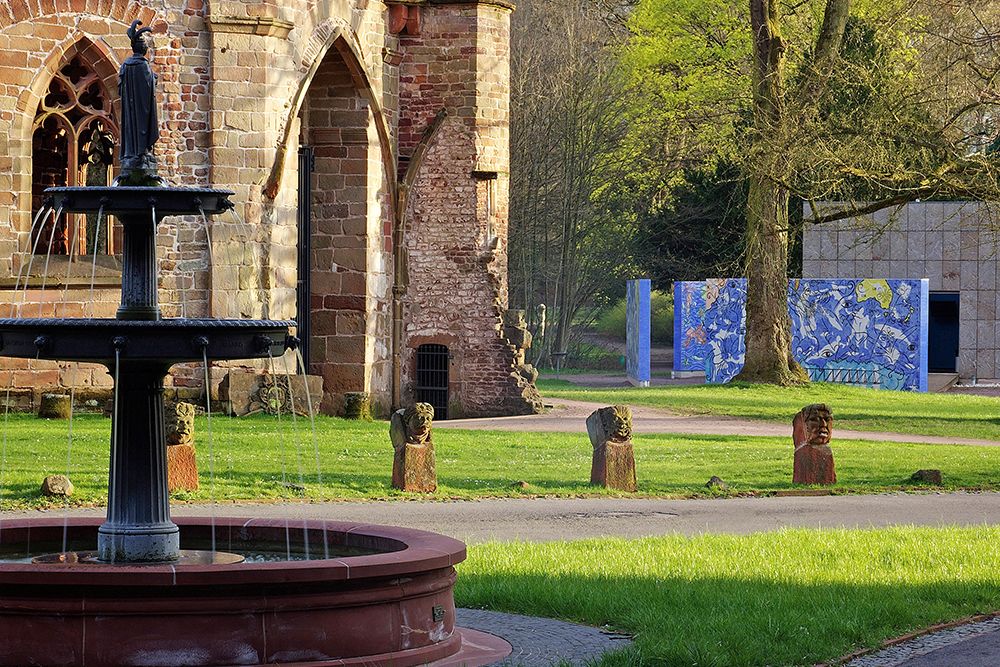Stone fountain and historic building in the park with sculptures and blue mural in the background.