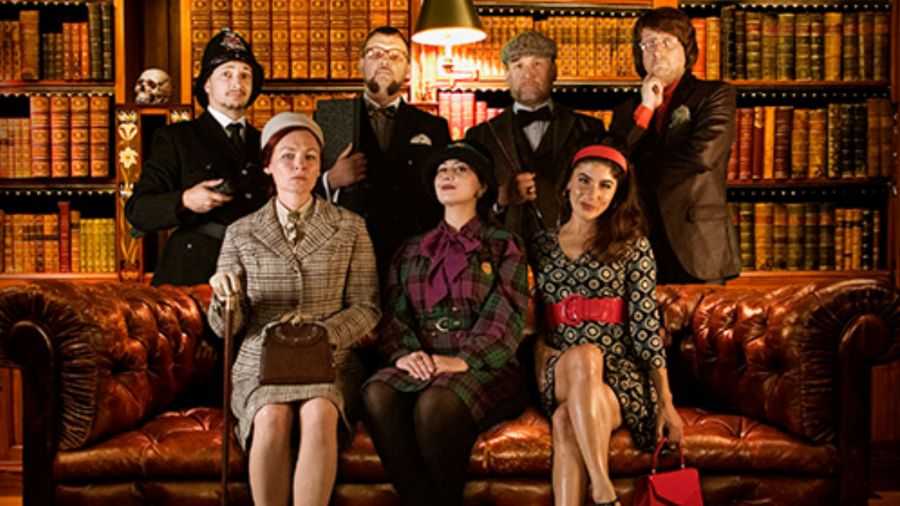 Sieben Personen posieren in einer Bibliothek mit ledernen Sofas und Bücherregalen. Drei sitzen auf einem Sofa, vier stehen dahinter. Alle tragen Kleidung im Retro-Stil.