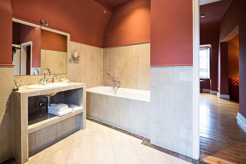 Badezimmer mit beige gefliester Badewanne, Waschbecken und roten Wänden, mit Spiegel und Handtüchern auf dem offenen Regal unter dem Waschbecken.