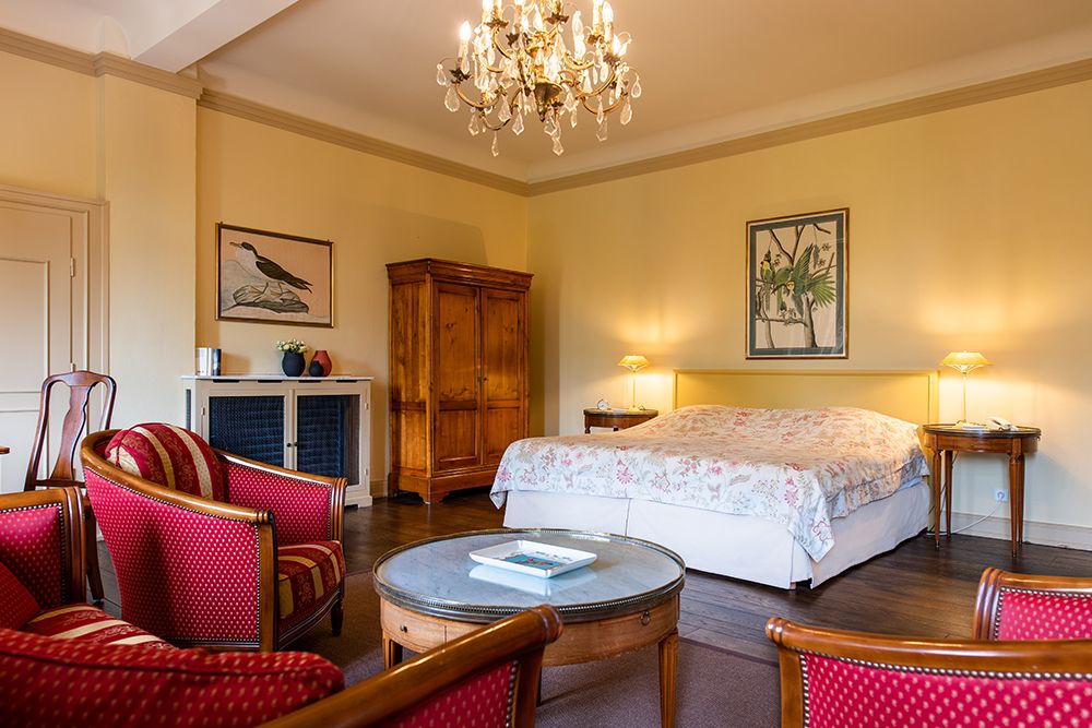Helles Schlafzimmer mit Kronleuchter, Holzschrank, Doppelbett mit geblümter Bettwäsche, roten Sesseln und gerahmter Kunst an den Wänden.