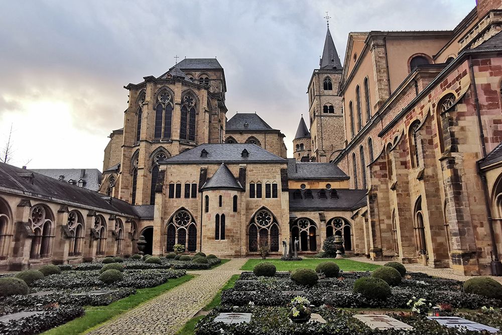 Entdecken Sie die majestätische, historische Steinkathedrale mit gotischer Architektur, Bögen und Türmen in Trier. Spazieren Sie bei einer inspirierenden Stadttour unter bewölktem Himmel durch den angelegten Innenhof mit symmetrischen Gärten und einem Steinpfad.