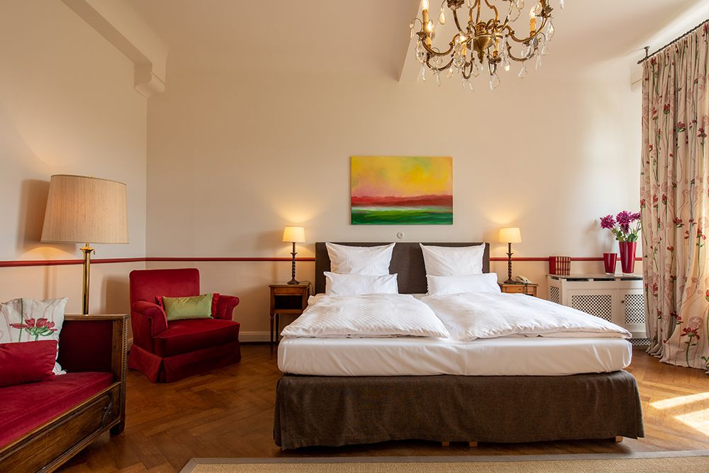 Elegantes Schlafzimmer mit Doppelbett, rotem Sessel, Kronleuchter, Lampen und farbenfrohen Kunstwerken sowie geblümten Vorhängen.