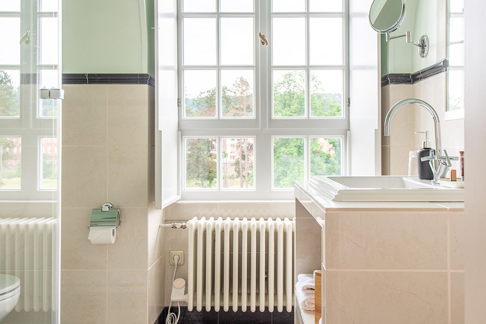 Helles Badezimmer mit großen Fenstern, weißem Waschbecken und Heizkörper darunter. Grüne und cremefarbene Wände, Spiegel und Toilettenartikel sichtbar.