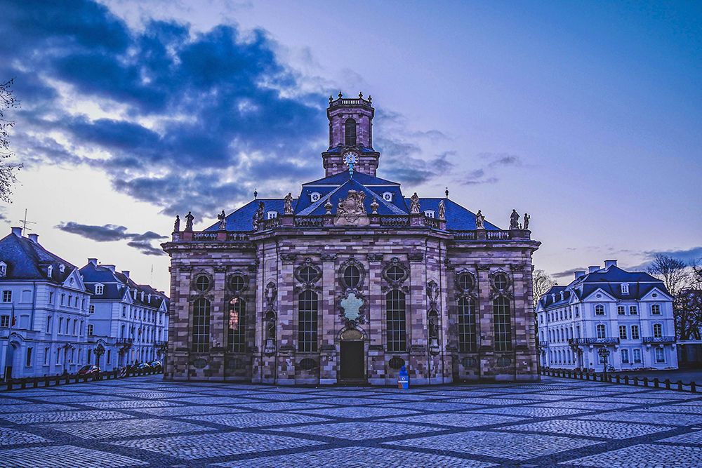 Kirche im Barockstil mit verzierter Fassade und Turm vor einem Abendhimmel mit vereinzelten Wolken in Saarbrücken. Offener, gepflasterter Platz im Vordergrund, perfekt für einen Abendspaziergang nach dem Einkaufen, mit umliegenden Gebäuden.