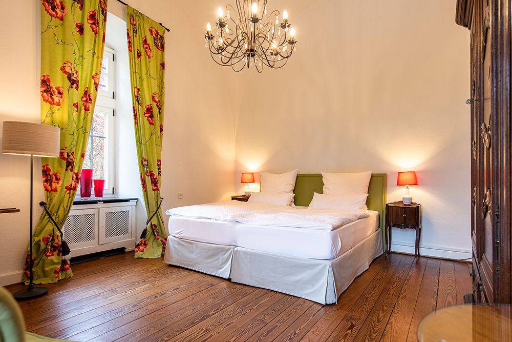 Schlafzimmer mit Doppelbett, geblümten Vorhängen, dekorativem Kronleuchter, Holzboden, zwei Nachttischen mit Lampen und einem großen Kleiderschrank.