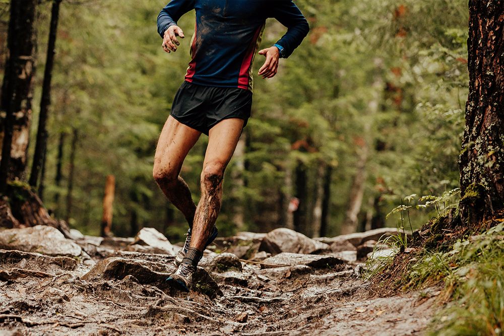 A person is jogging on a muddy, rocky path in the woods. He is wearing a long-sleeved shirt and shorts and his legs are splattered with mud.