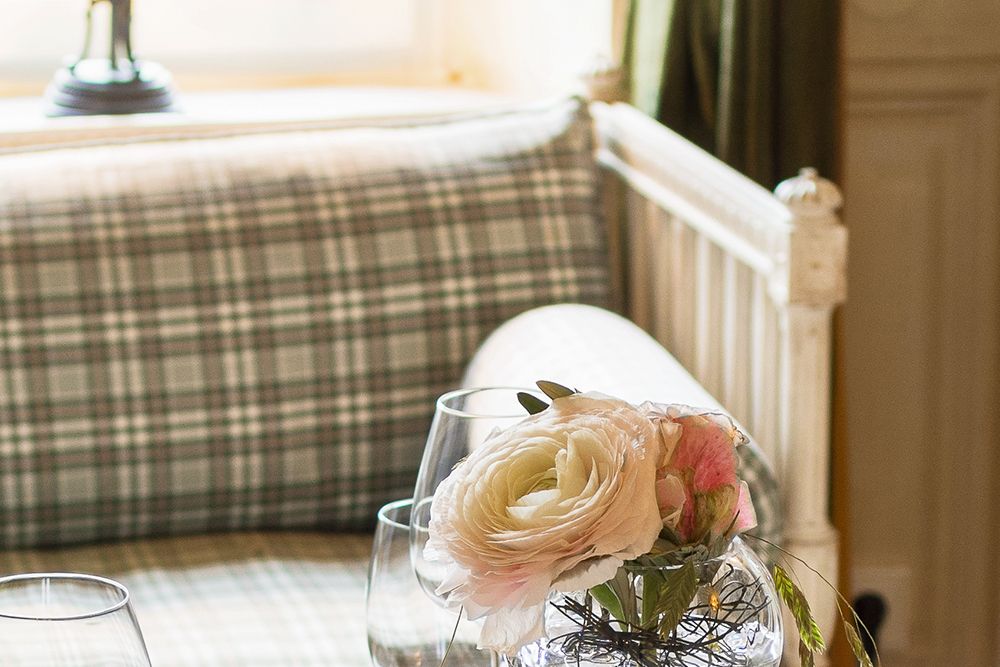 Ein kleiner Strauß rosafarbener und weißer Blumen in einer Vase ziert den für ein gemütliches Abendessen zu zweit gedeckten Tisch. Vor einem karierten Polstersofa am Fenster funkeln Weingläser.