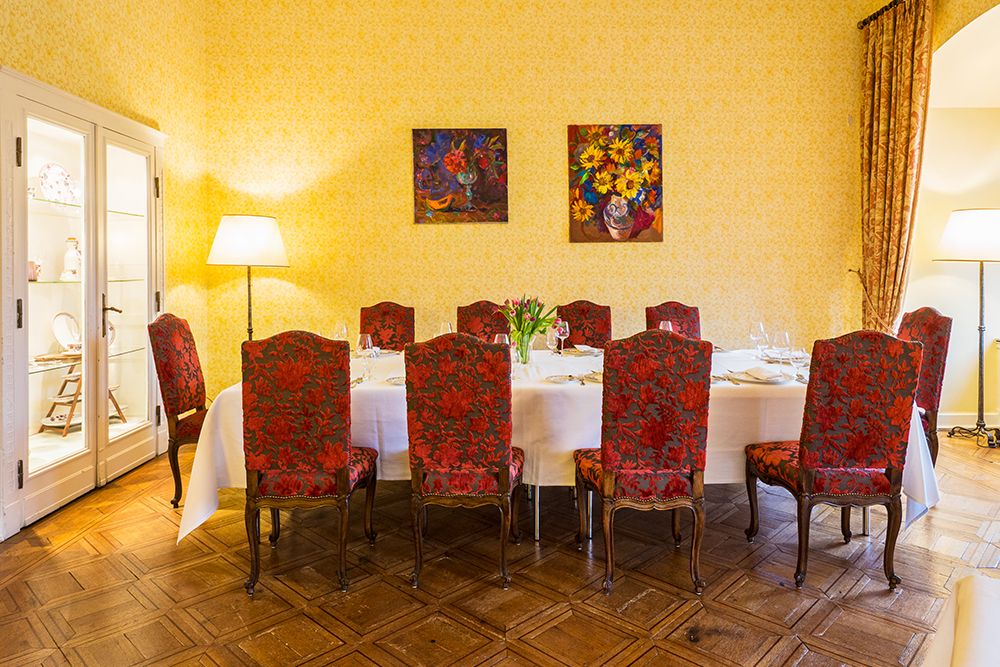 Elegant dining room with a large table for eight people, perfect for celebrations. Red upholstered chairs surround the table covered in white fabric. Yellow patterned walls, reminiscent of Anna Boch's style, feature two floral paintings.