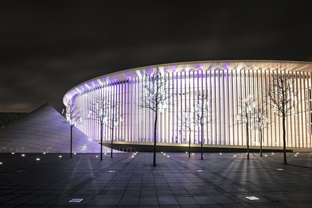 Eine Nachtansicht des modernen Kultur-Gebäudes zeigt vertikale Säulen, die in violettes Licht getaucht sind. Im Vordergrund verstärken kahle Bäume und eine kleine Pyramidenstruktur den Reiz der Szene.
