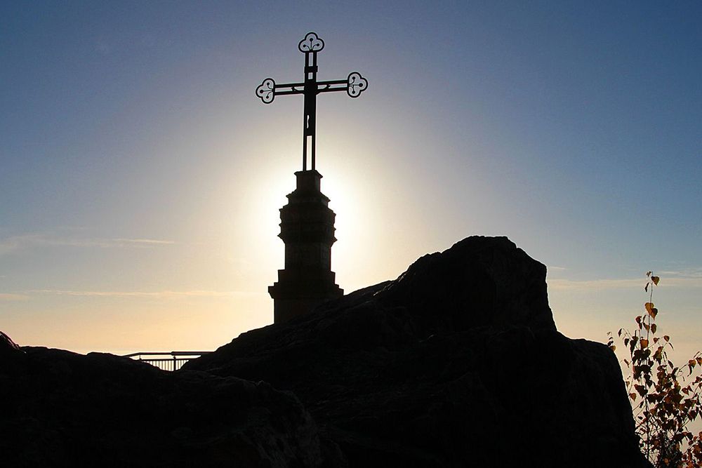 Silhouette of a cross on a hill, backlit by the sun, surrounded by rocks and sparse vegetation.