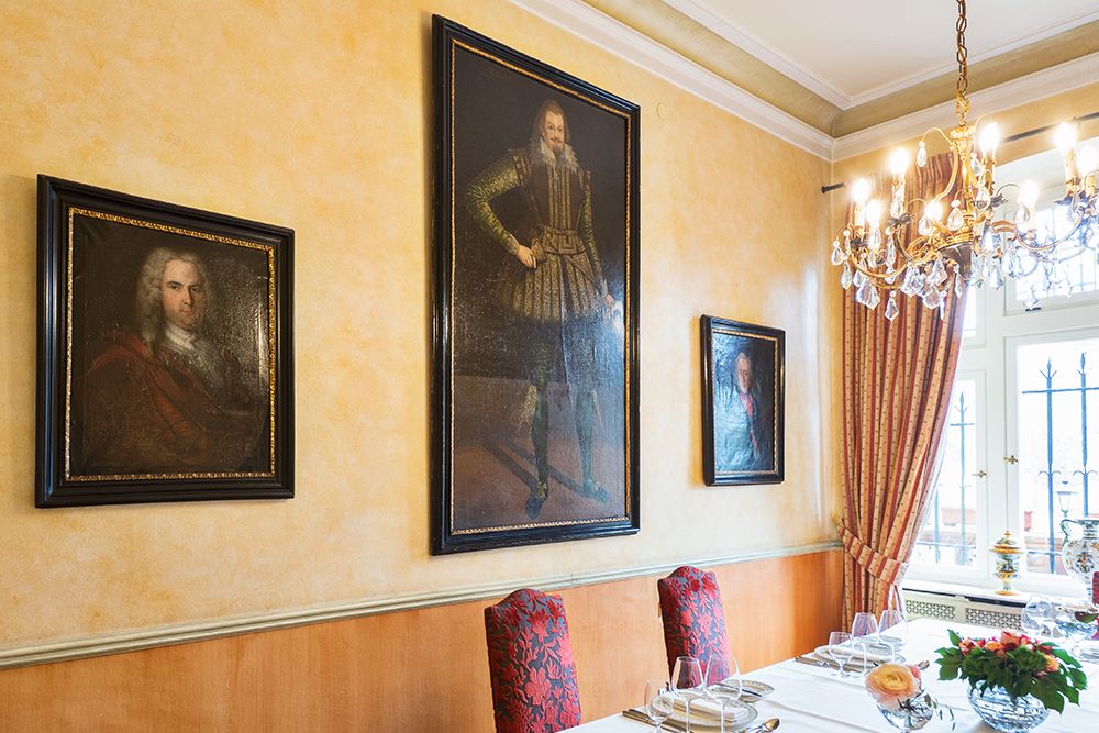 Der Speisesaal strahlt mit seinen verzierten Stühlen und dem gedeckten Tisch eine stimmungsvolle Atmosphäre aus. Drei historische Porträts zieren die gelben Wände, die an Alt Straßbourg erinnern. Darüber leuchtet ein Kronleuchter, während die großen Fenster rechts mit eleganten Vorhängen geschmückt sind.