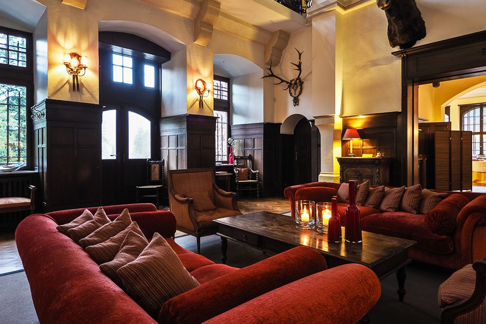 Ein Raum mit roten Sofas, einem hölzernen Couchtisch und an der Wand montierten Geweihen strahlt einen gemütlichen Charme aus. Große Fenster mit gewölbten Oberlichtern lassen Licht herein, während Kerzen und Lampen das gemütliche, rustikale Ambiente verstärken, das an eine einladende Hotellobby erinnert, die sich perfekt für einen Aperitif eignet.