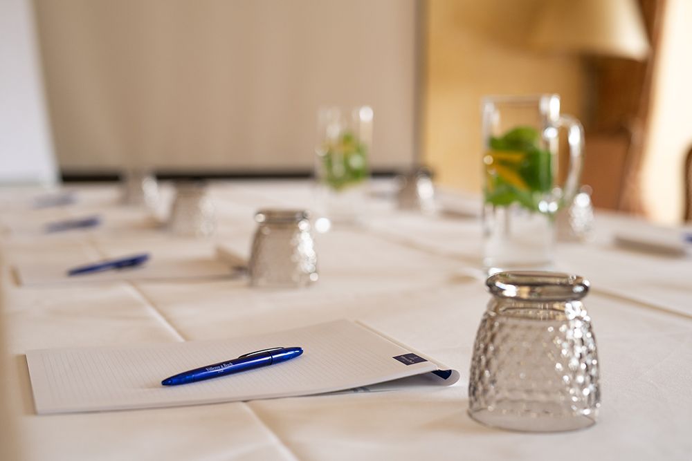 Nahaufnahme eines Tagungskonferenztischs mit Notizblöcken, Stiften, umgedrehten Gläsern und einer Kanne mit aromatisiertem Wasser, alles sorgfältig angeordnet in gut ausgestatteten Räumlichkeiten.