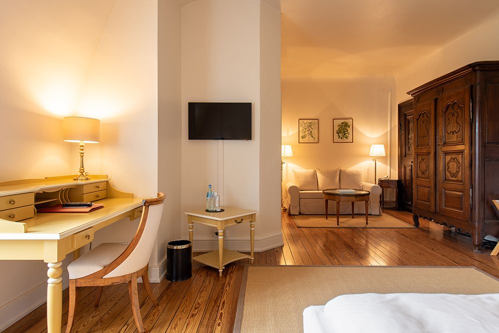 Geräumiges Zimmer mit gelbem Schreibtisch, Stuhl, Wandfernseher, Sofa und großem Holzschrank. Lampen erhellen den Raum sanft.