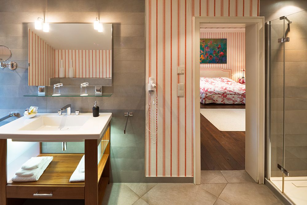 Modernes Badezimmer mit Waschbecken, Spiegel und Glasdusche. Durch die Tür gelangt man ins Schlafzimmer mit gemusterter Tagesdecke und farbenfrohen Kunstwerken.