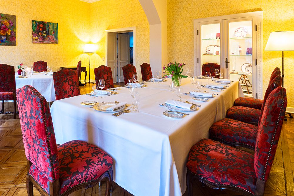 Elegant dining room with a long white table set with glasses and cutlery. Red upholstered chairs surround the table. Warm lighting and art by Anna Boch decorate the yellow walls, perfect for meetings or small celebrations.