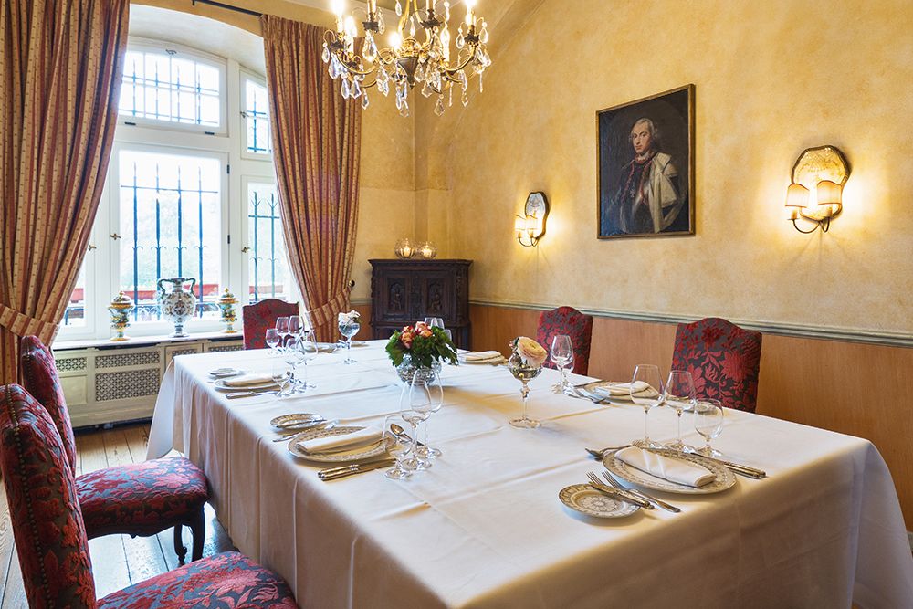 Dieser elegante Speisesaal strahlt eine stimmungsvolle Atmosphäre aus, perfekt für Feiern. Ein langer, für das Abendessen gedeckter Tisch, umgeben von roten Polsterstühlen, steht unter einem Kronleuchter. Ein Porträt ziert die Wand, während natürliches Licht durch die hohen Fenster fällt.