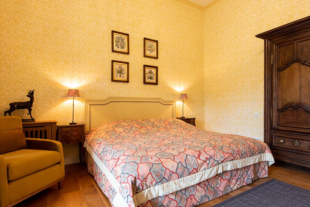 Schlafzimmer mit großem Bett, gemusterter Tagesdecke, zwei Beistelltischen mit Lampen, gelbem Sessel und gerahmten Bildern an gelben Wänden.