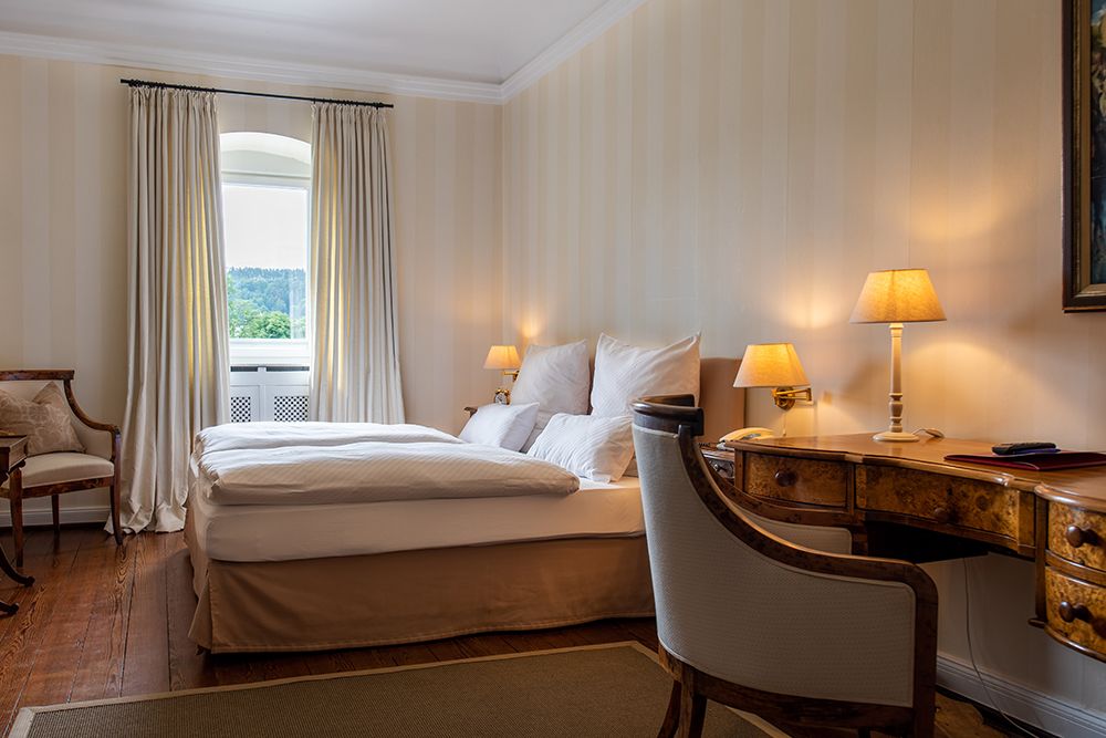 Hotelzimmer mit Doppelbett, Holzschreibtisch, Stühlen und Lampen. Ein Fenster mit Vorhängen bietet einen Blick ins Grüne.