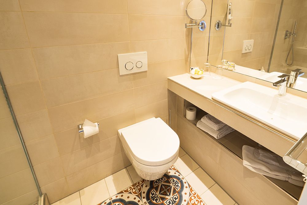 Ein modernes Badezimmer mit einer wandmontierten Toilette, einem Waschbecken mit Spiegel und einem beigen Fliesendesign mit gemusterten Bodenfliesen.