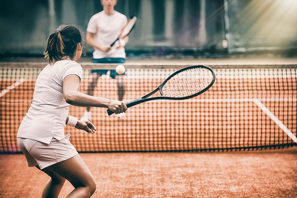 A woman and a man are engaged in an exciting tennis match on a clay court. Her concentration is palpable as she prepares to hit the ball. Both are dressed in crisp white, embodying the classic elegance of tennis as they enjoy this energetic mix of jogging and sport.