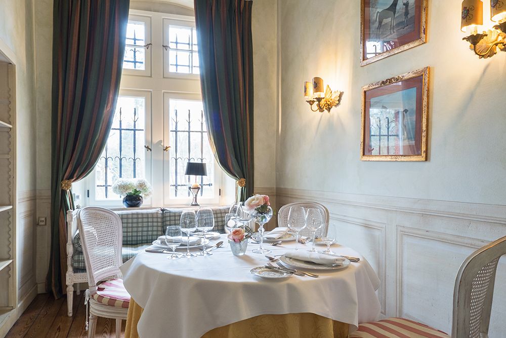 Elegantes Esszimmer mit einem runden Tisch für ein gemütliches Abendessen zu zweit, mit weißen Tellern, Gläsern und einem Blumenschmuck. Hohe Fenster mit Vorhängen sorgen für Privatsphäre, ergänzt durch gerahmte Kunstwerke an hellen Wänden.