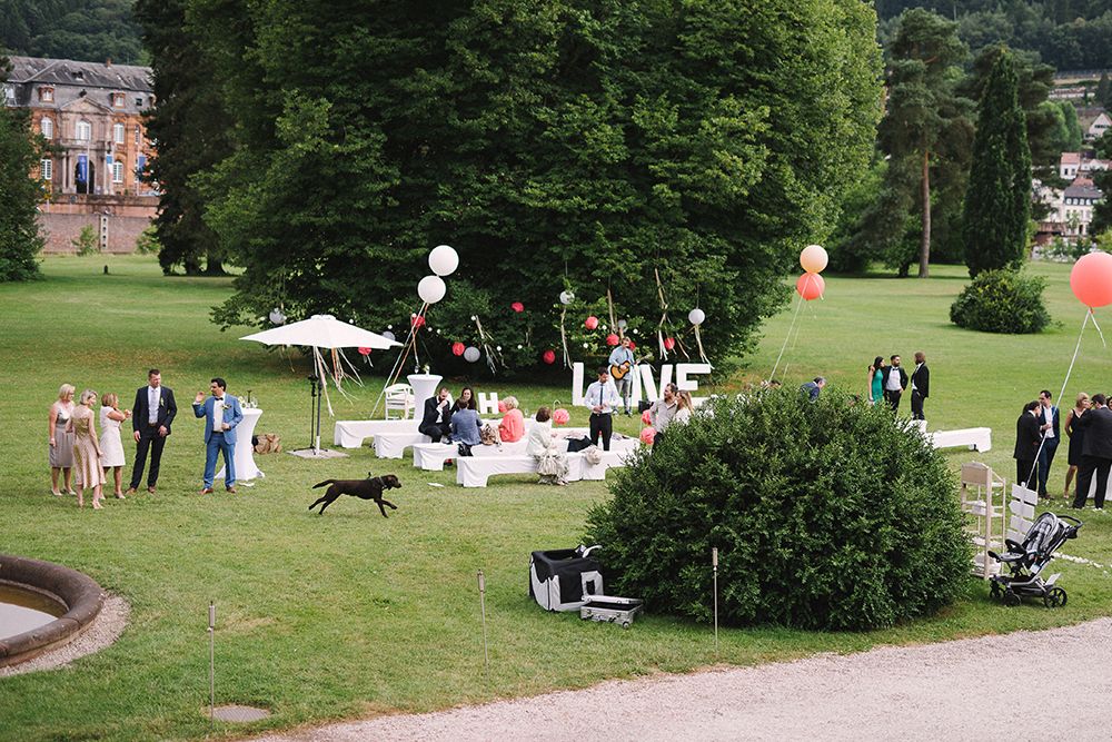 Outdoor wedding scene with guests milling around on a grassy area. Large "LOVE" letters, balloons and white seating are present. A dog runs across the grass. Trees and a building are visible in the background.