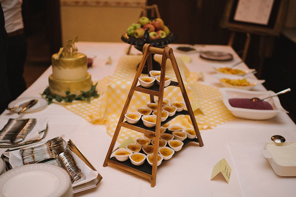 Dessert table with a layered wooden stand holding small cups, a golden cake, a bowl of fruit, cutlery, plates and a bowl of purple dessert on a yellow tablecloth.