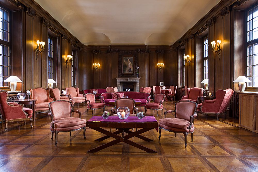 Ein elegantes Kaminzimmer mit holzgetäfelten Wänden, großen Fenstern und Vintage-Möbeln, darunter symmetrisch angeordnete rote Polsterstühle und Sofas. In der Mitte der Rückwand befindet sich ein Kamin, der sich perfekt zum Genießen eines feinen Single Malt Whiskeys eignet.