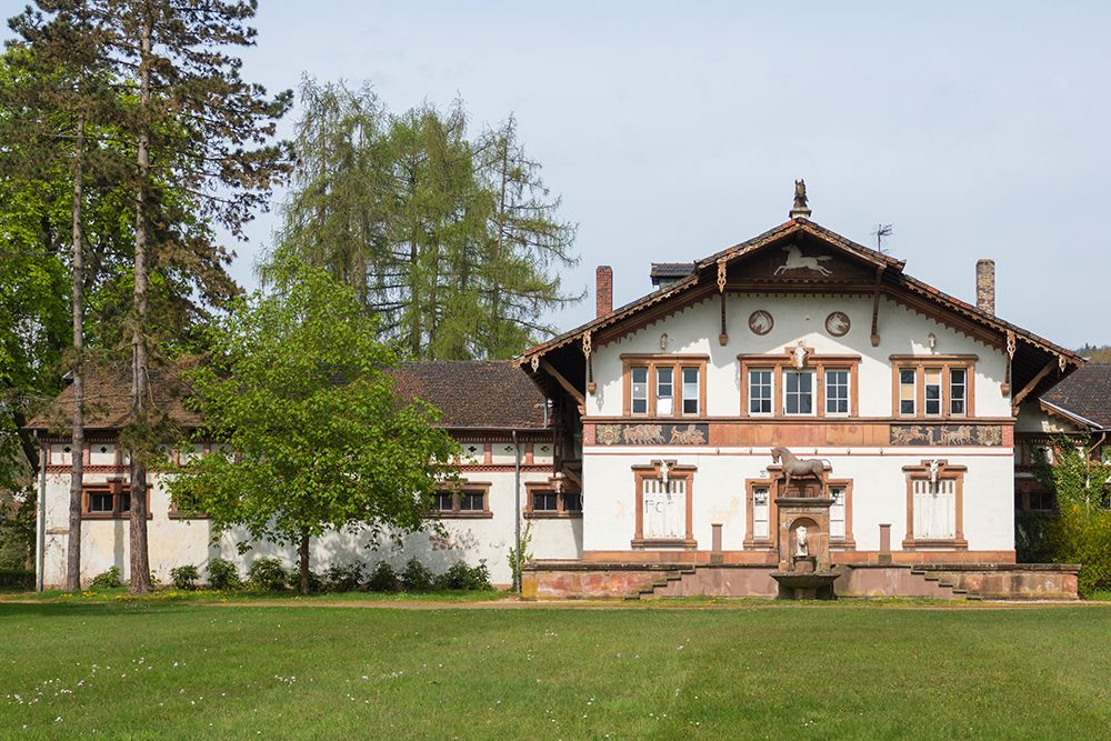 Ein historisches Gebäude mit einer Fassade aus Holz und Stein und einem Brunnen in der Mitte, umgeben von Bäumen und an klaren Tagen von einer Wiese.