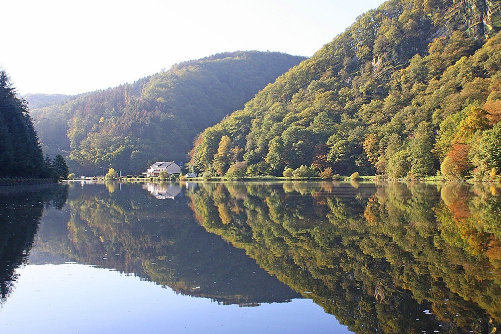 A calm lake reflects wooded hills and a distant building under a clear sky.