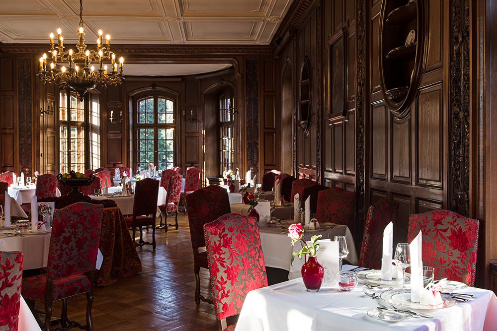 Elegantes Esszimmer mit kunstvoller Holzvertäfelung, rot gemusterten Stühlen, weißen Tischdecken und einem Kronleuchter. Perfekt für festliche Hochzeiten, die Tische sind mit Weingläsern und Servietten gedeckt, während große Fenster natürliches Licht hereinlassen.