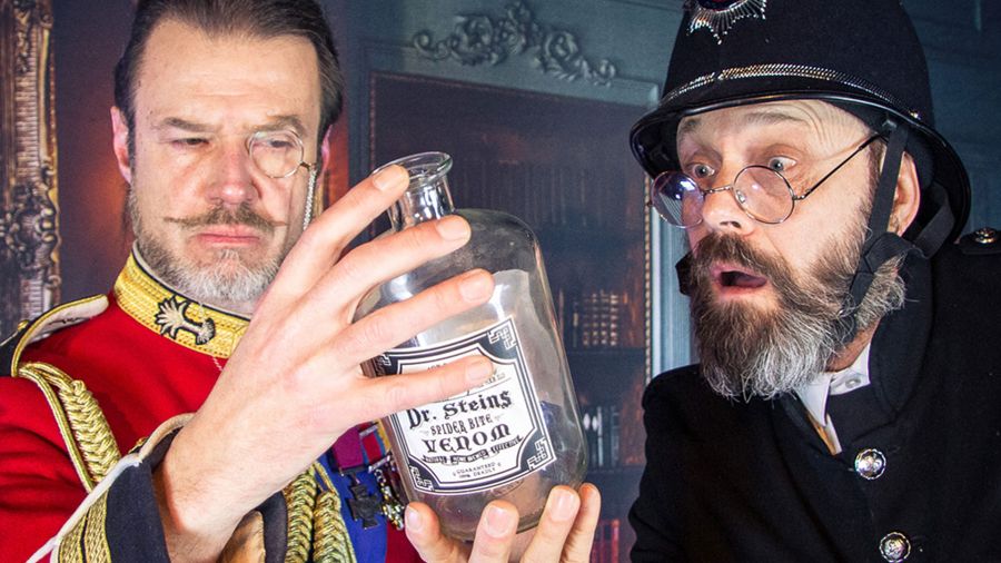 Zwei Männer in Vintage-Kostümen untersuchen in einer Theaterkulisse eine Glasflasche mit der Aufschrift „Dr. Stein’s Stage Use Venom“.