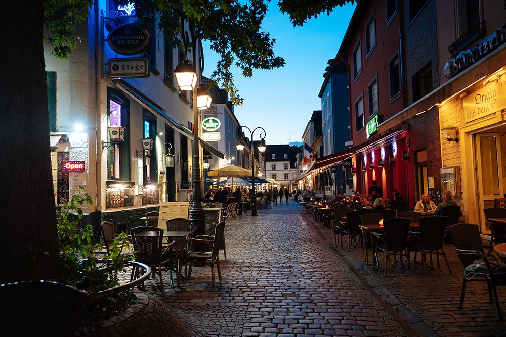 Cobblestone street with outdoor seating on either side of a café, illuminated streetlights and people dining under an evening sky.