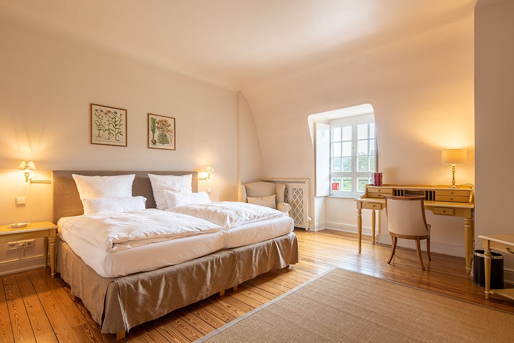 Hotelzimmer mit zwei Betten, Schreibtisch, Stuhl und Fenster. Die Wände sind mit gerahmten Kunstwerken geschmückt. Warme Beleuchtung und Holzboden.