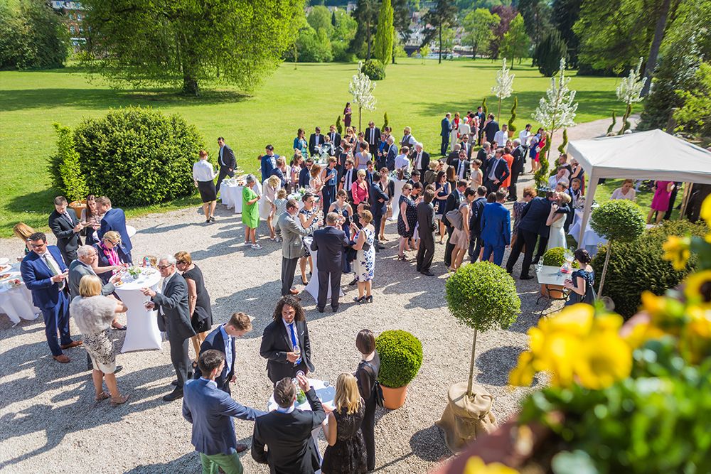 A large group of people in formal attire gather outdoors at a garden event with tables and decorations.